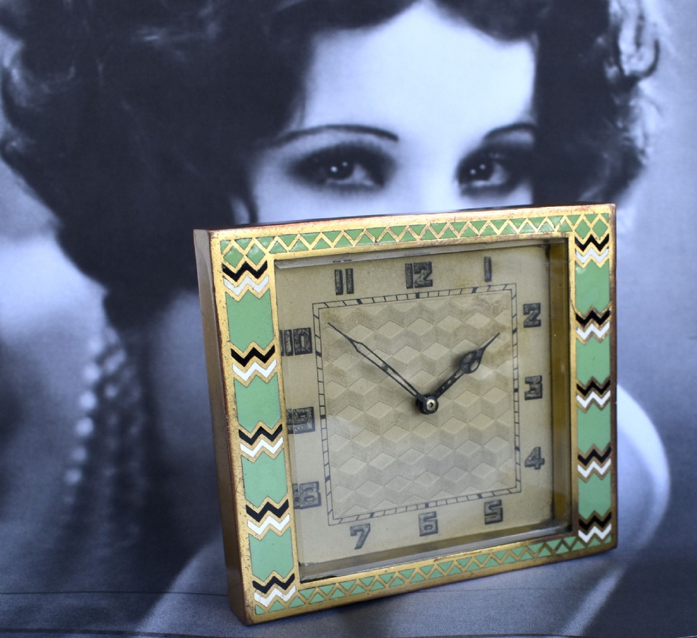 A square Art Deco clock featuring a green, white, and black chevron enamel border surrounding a geometric patterned metal dial with Arabic numerals. View VI