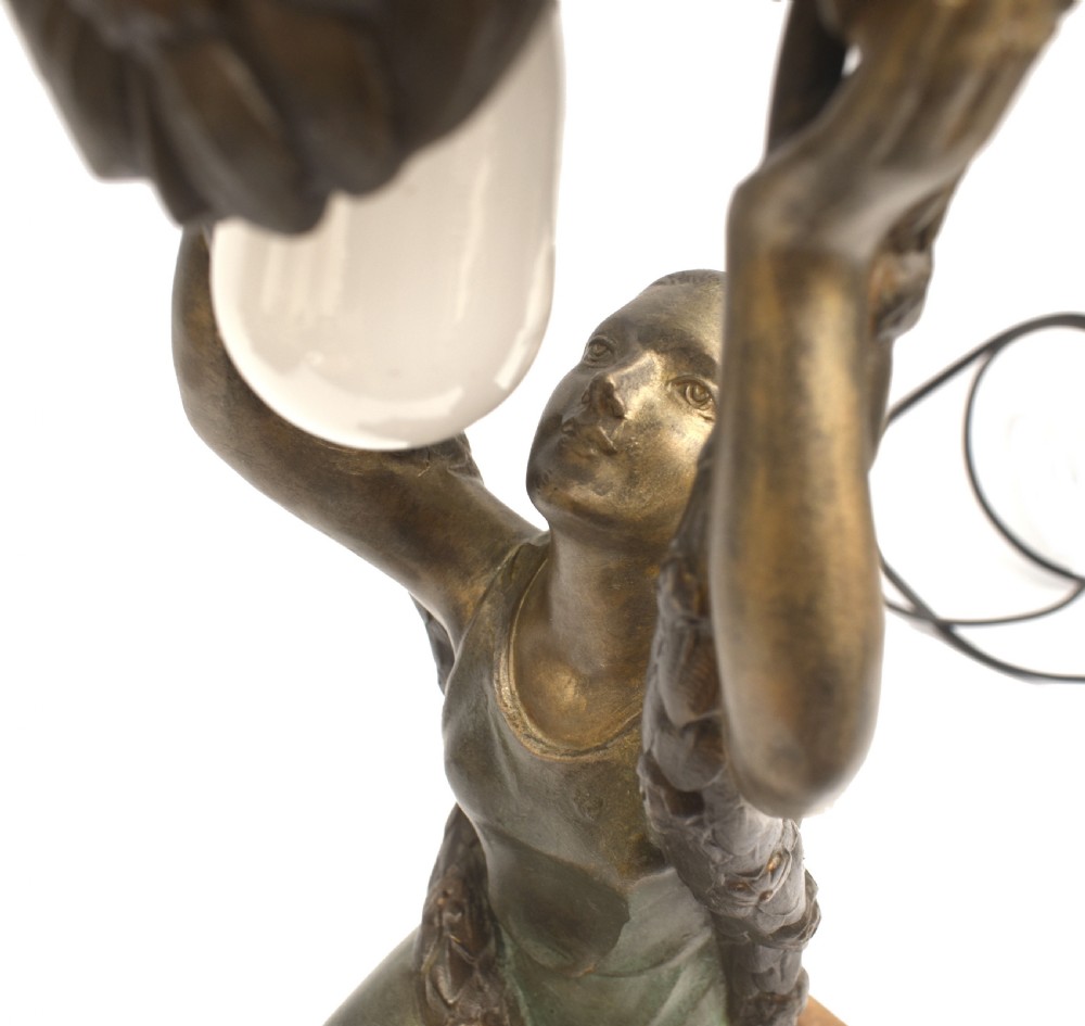 An Art Deco patinated metal table lamp featuring a kneeling female figure holding up a floral garland shade, set on a rectangular marble base. View X