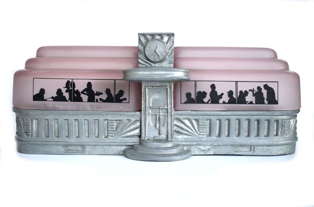 An Art Deco style lamp featuring a stepped pink frosted glass body decorated with black silhouetted figures dining inside windows, framed by a silver-coloured cast metal base and central architectural doorway with a clock motif.