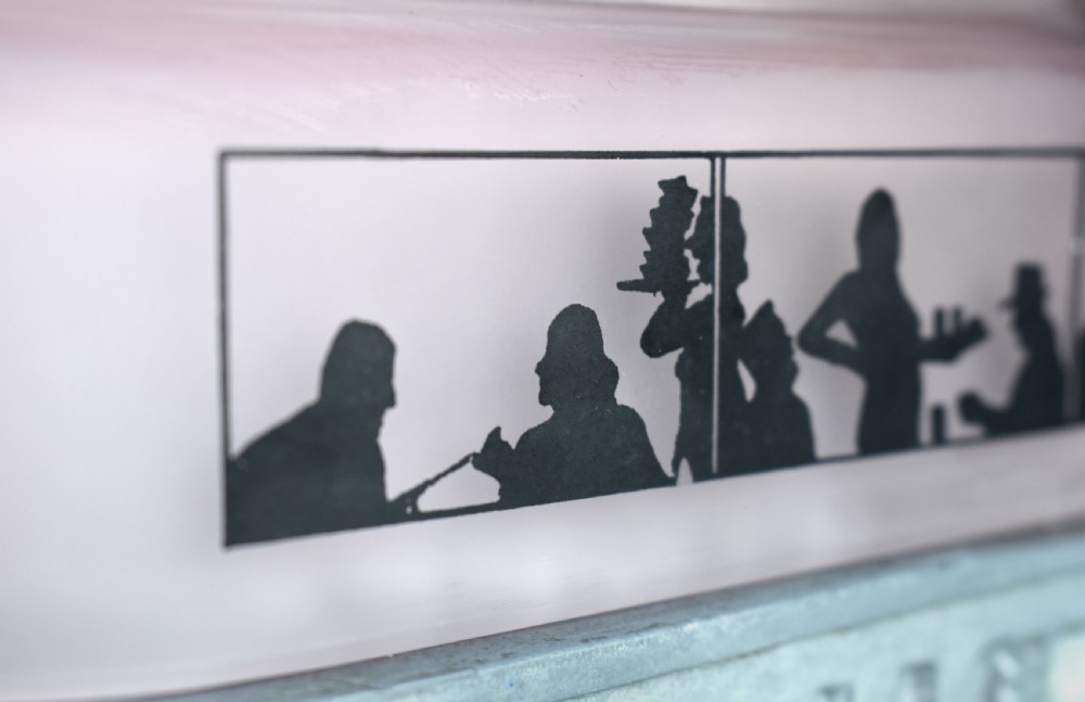 An Art Deco style lamp featuring a stepped pink frosted glass body decorated with black silhouetted figures dining inside windows, framed by a silver-coloured cast metal base and central architectural doorway with a clock motif. View VI