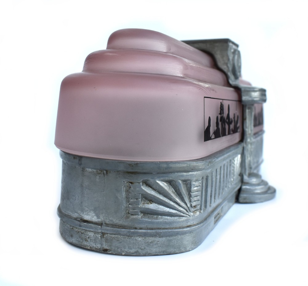 An Art Deco style lamp featuring a stepped pink frosted glass body decorated with black silhouetted figures dining inside windows, framed by a silver-coloured cast metal base and central architectural doorway with a clock motif. View X