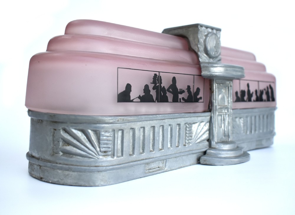 An Art Deco style lamp featuring a stepped pink frosted glass body decorated with black silhouetted figures dining inside windows, framed by a silver-coloured cast metal base and central architectural doorway with a clock motif. View XI