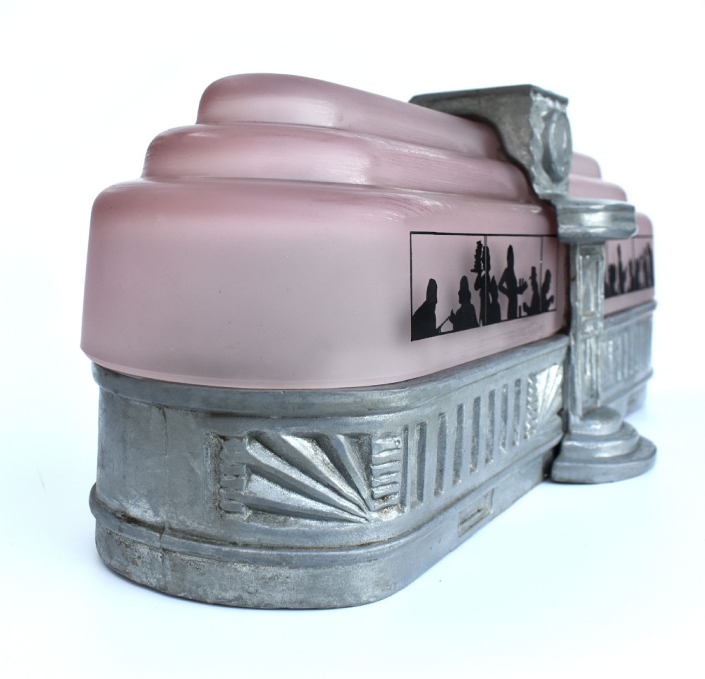 An Art Deco style lamp featuring a stepped pink frosted glass body decorated with black silhouetted figures dining inside windows, framed by a silver-coloured cast metal base and central architectural doorway with a clock motif. View XII