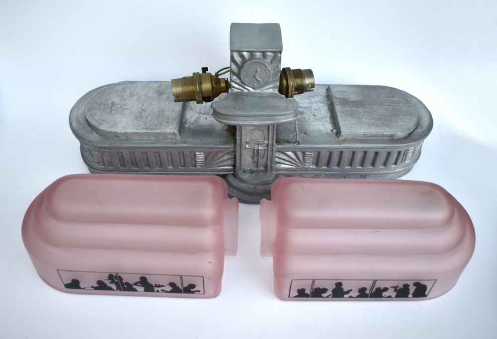 An Art Deco style lamp featuring a stepped pink frosted glass body decorated with black silhouetted figures dining inside windows, framed by a silver-coloured cast metal base and central architectural doorway with a clock motif. View XIII