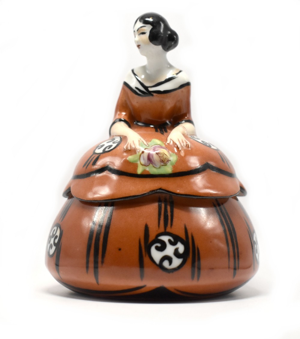 An Art Deco glazed ceramic powder bowl featuring a lid shaped as a lady in a terracotta and black dress holding a flower.