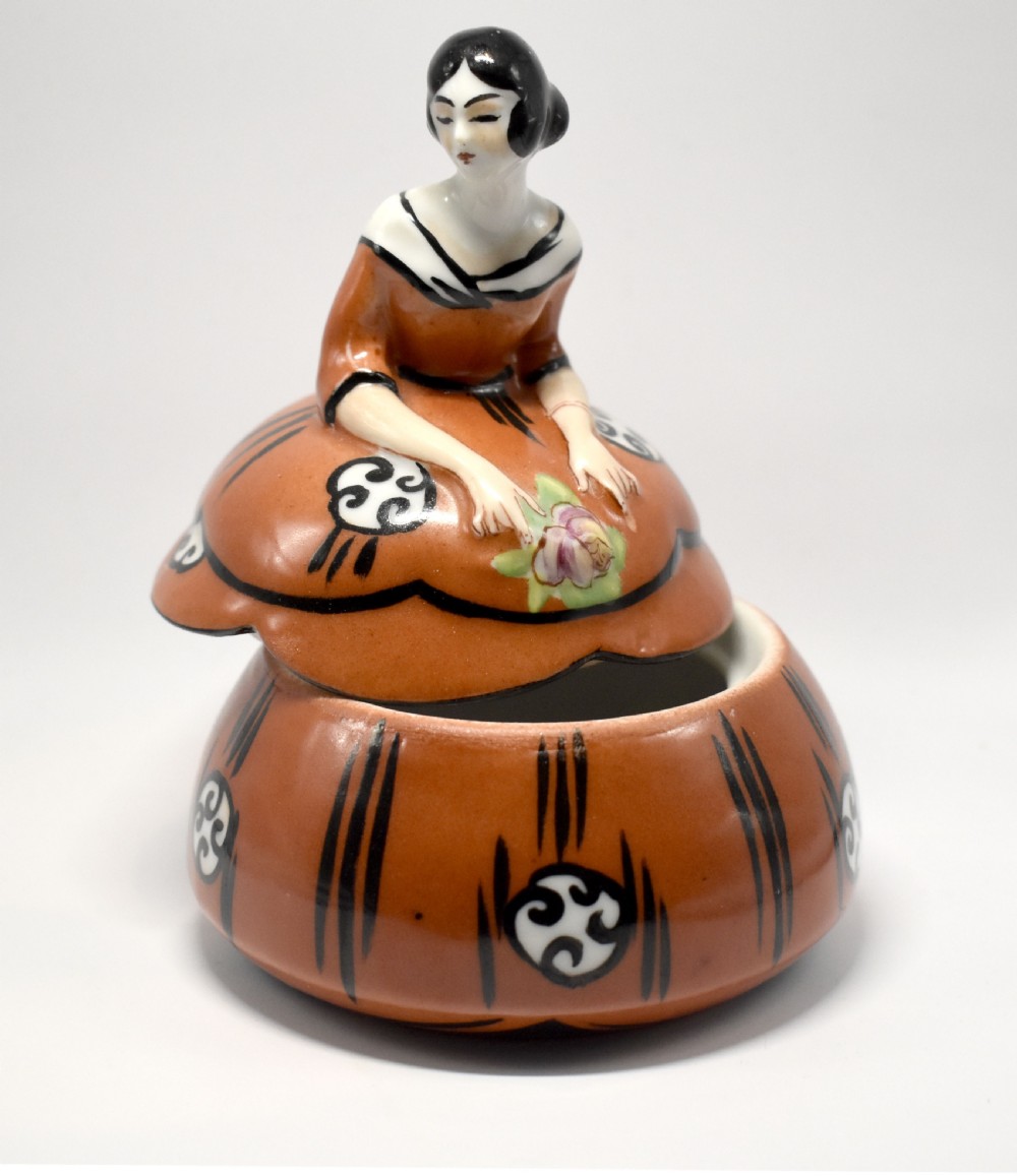 An Art Deco glazed ceramic powder bowl featuring a lid shaped as a lady in a terracotta and black dress holding a flower. View II