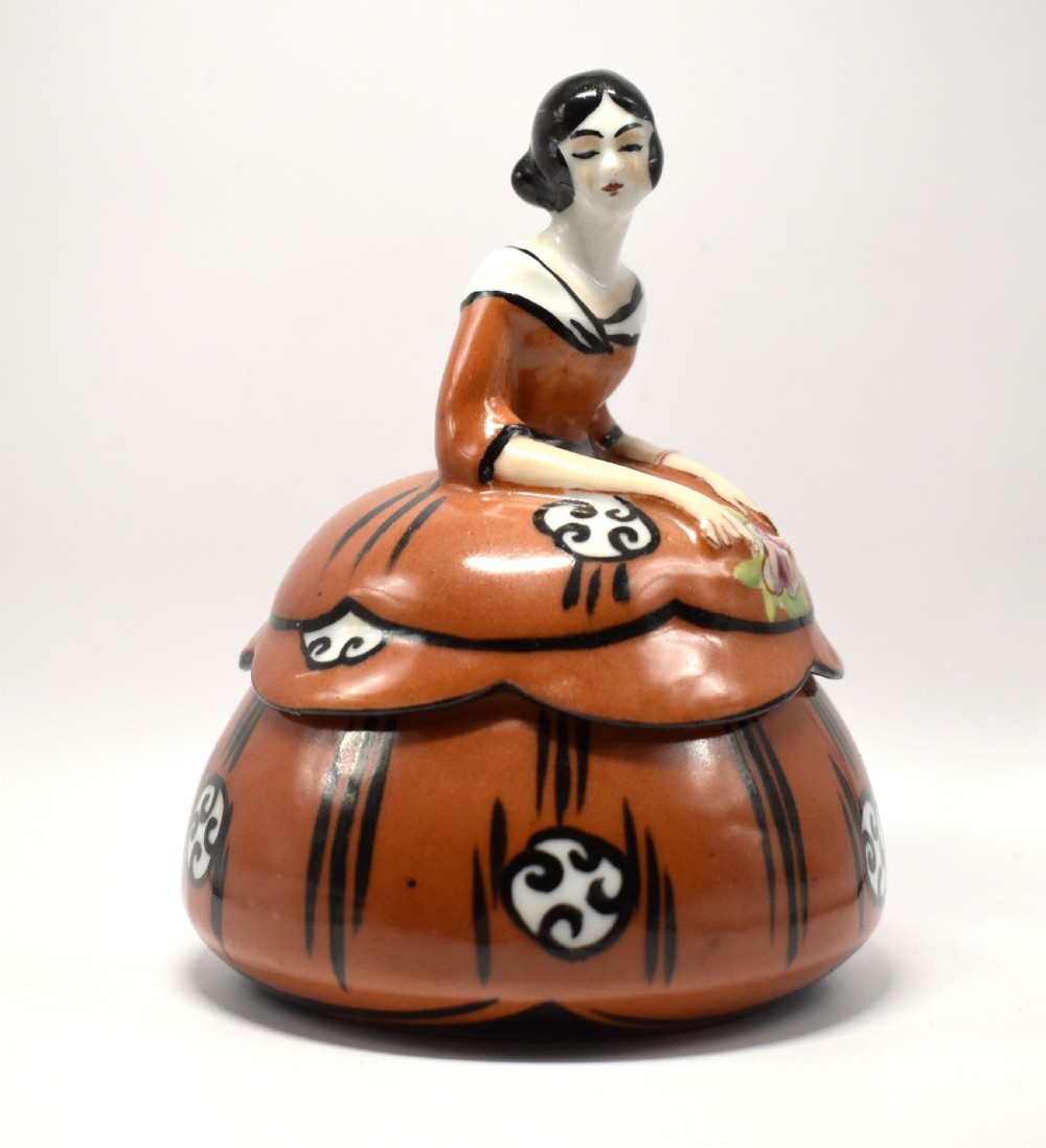 An Art Deco glazed ceramic powder bowl featuring a lid shaped as a lady in a terracotta and black dress holding a flower. View V