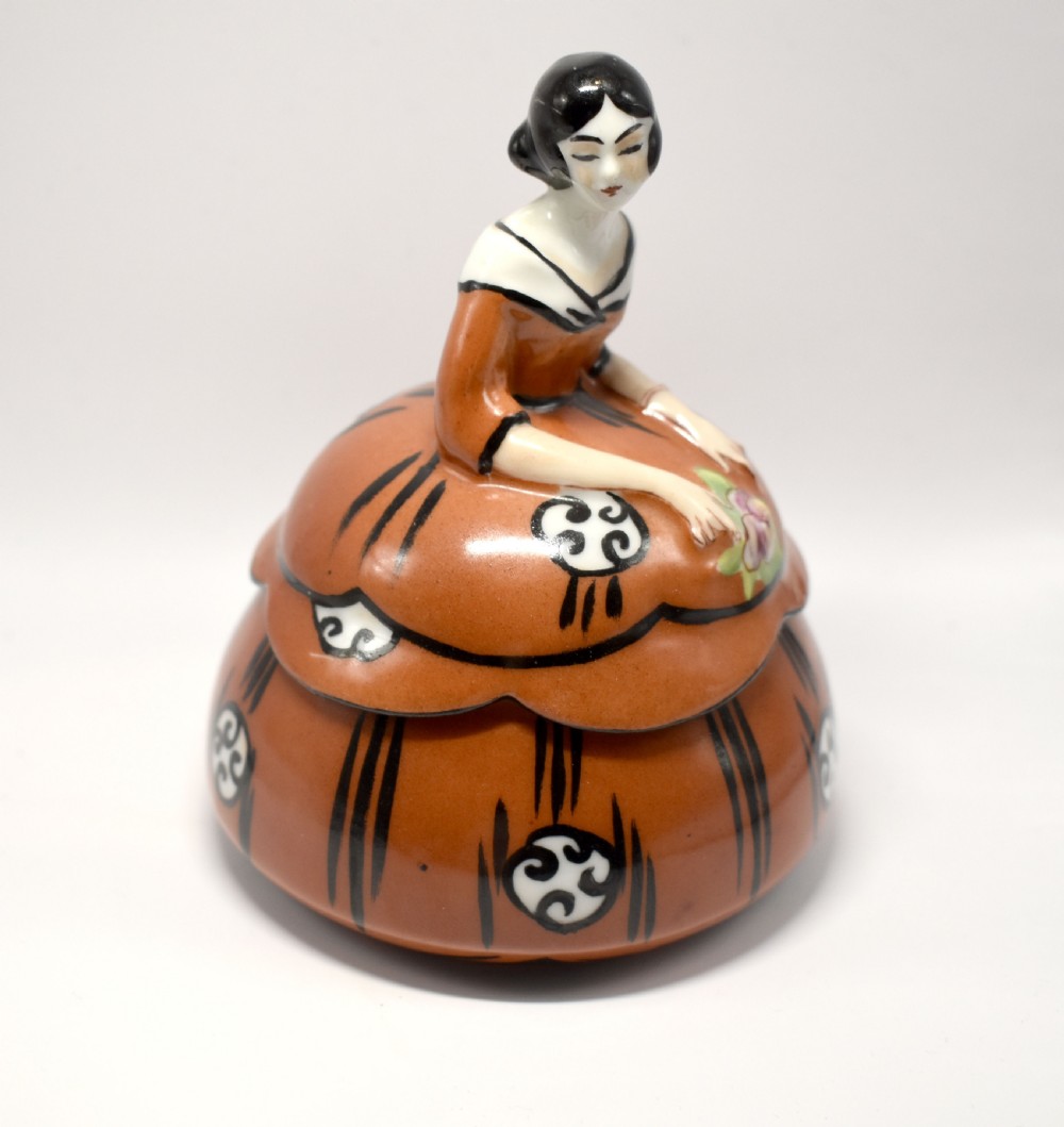 An Art Deco glazed ceramic powder bowl featuring a lid shaped as a lady in a terracotta and black dress holding a flower. View VI