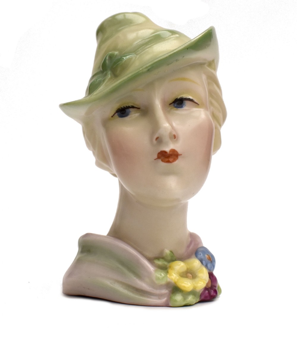 An Art Deco ceramic bust of a lady wearing a pale green hat and a floral corsage on a plain white background.