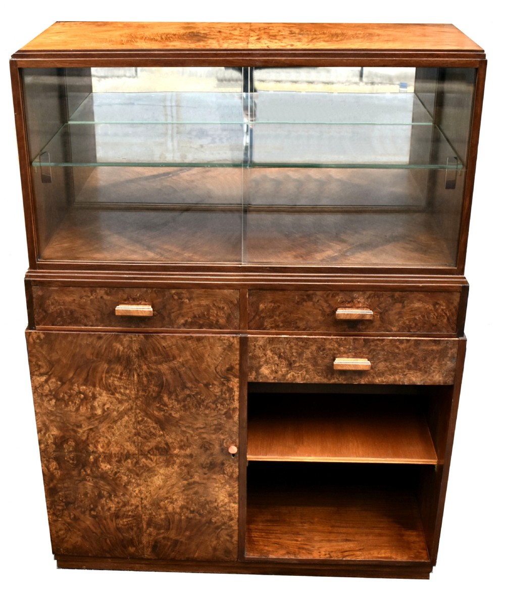 An Art Deco figured walnut drinks cabinet featuring a glass-fronted upper display section with glass shelves, drawers, and lower storage compartments.
