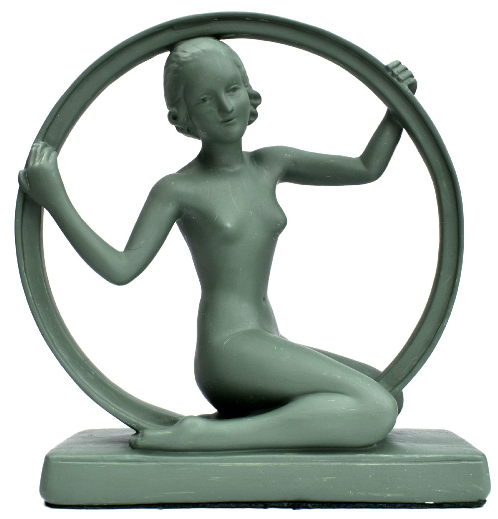 A sage green Art Deco painted plaster figurine depicting a kneeling female figure inside a circular hoop set on a rectangular base.