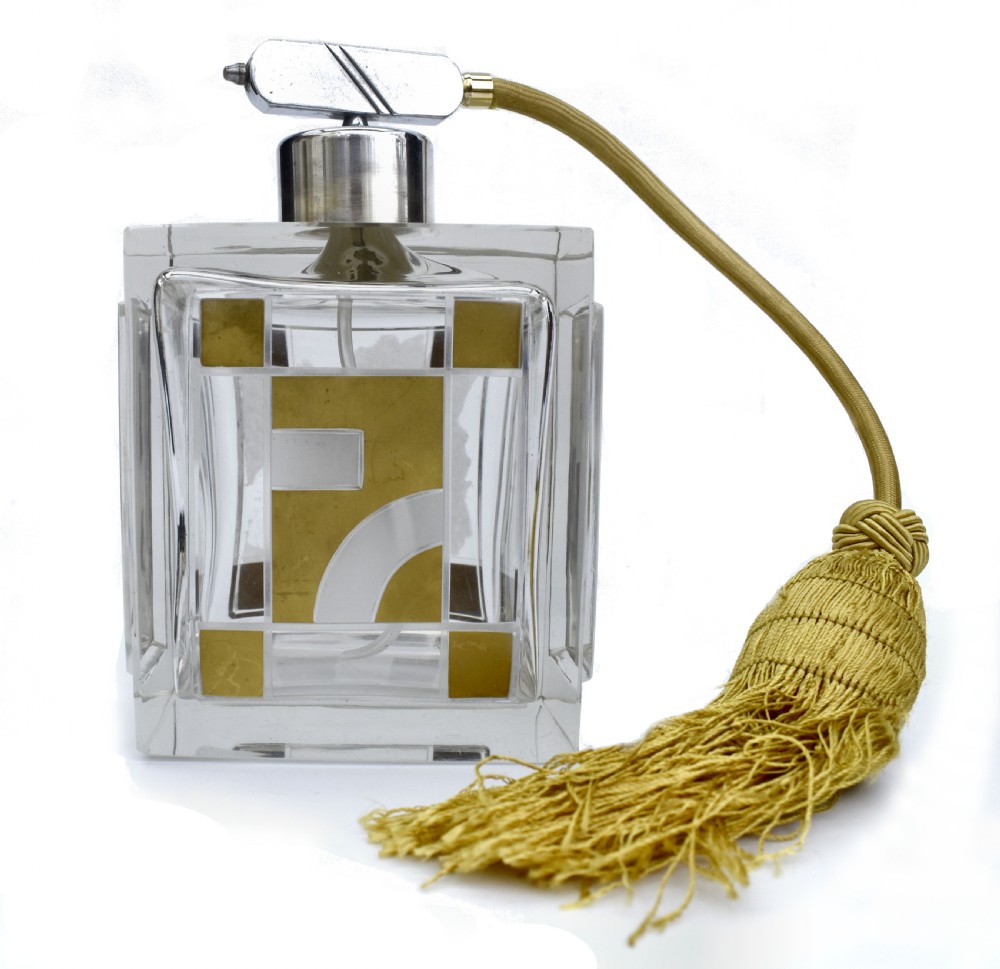 A square clear glass perfume atomizer featuring geometric gold decoration, a chrome pump mechanism, and a gold woven spray tube with a tassel. View IV