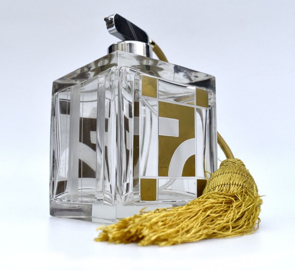 A square clear glass perfume atomizer featuring geometric gold decoration, a chrome pump mechanism, and a gold woven spray tube with a tassel.