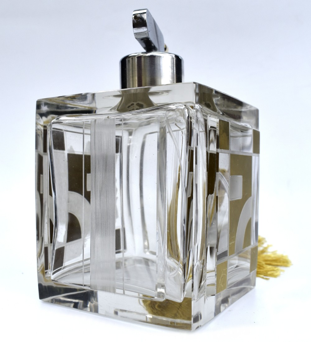 A square clear glass perfume atomizer featuring geometric gold decoration, a chrome pump mechanism, and a gold woven spray tube with a tassel. View VII