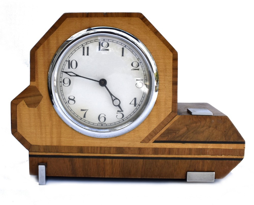 An asymmetrical Art Deco wooden mantel clock featuring two-tone wood veneers, a circular white dial with chrome bezel, and chrome foot accents.