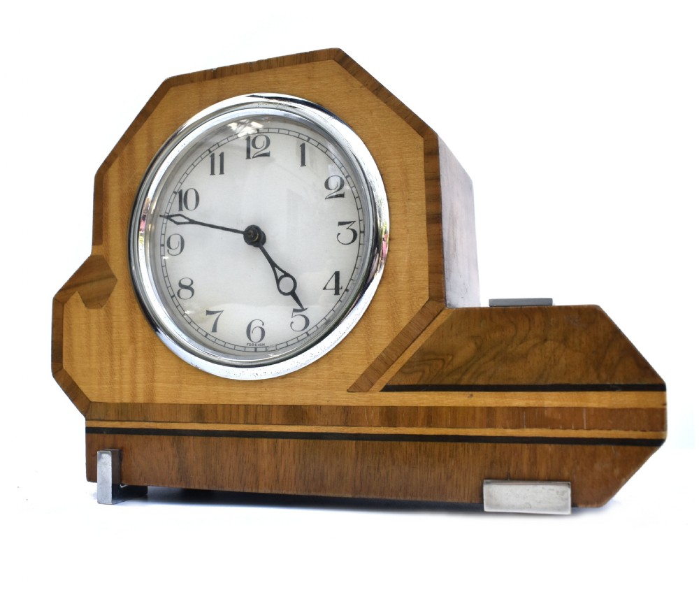An asymmetrical Art Deco wooden mantel clock featuring two-tone wood veneers, a circular white dial with chrome bezel, and chrome foot accents. View VI