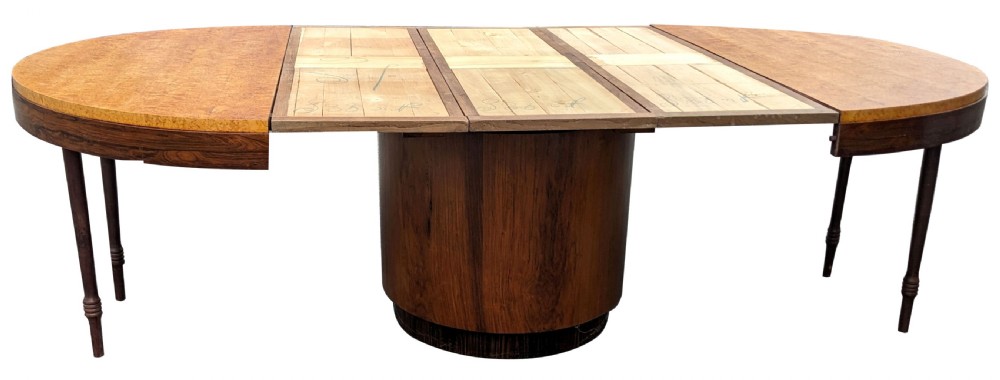 An extended Art Deco oval dining table featuring figured burr veneer ends, central pedestal base, extra insertion leaves, and slim support legs. View V