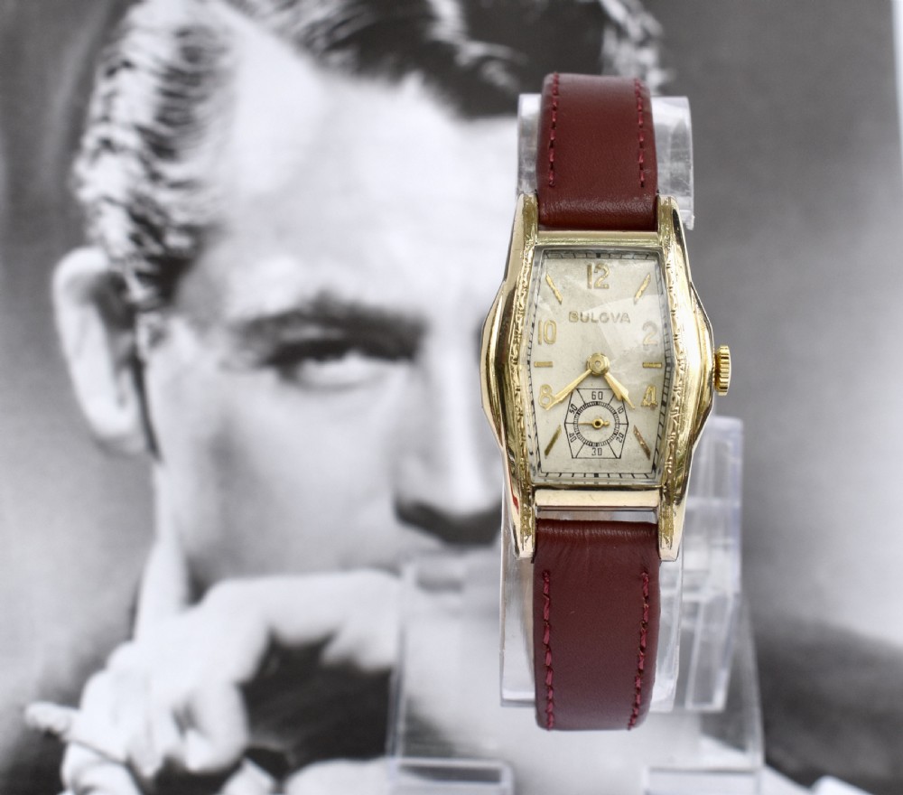 An Art Deco Bulova gents wristwatch featuring a gold-tone tonneau case with engraved side detailing, a cream dial with gold numerals, and a burgundy leather strap.
