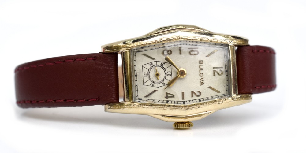 An Art Deco Bulova gents wristwatch featuring a gold-tone tonneau case with engraved side detailing, a cream dial with gold numerals, and a burgundy leather strap. View II