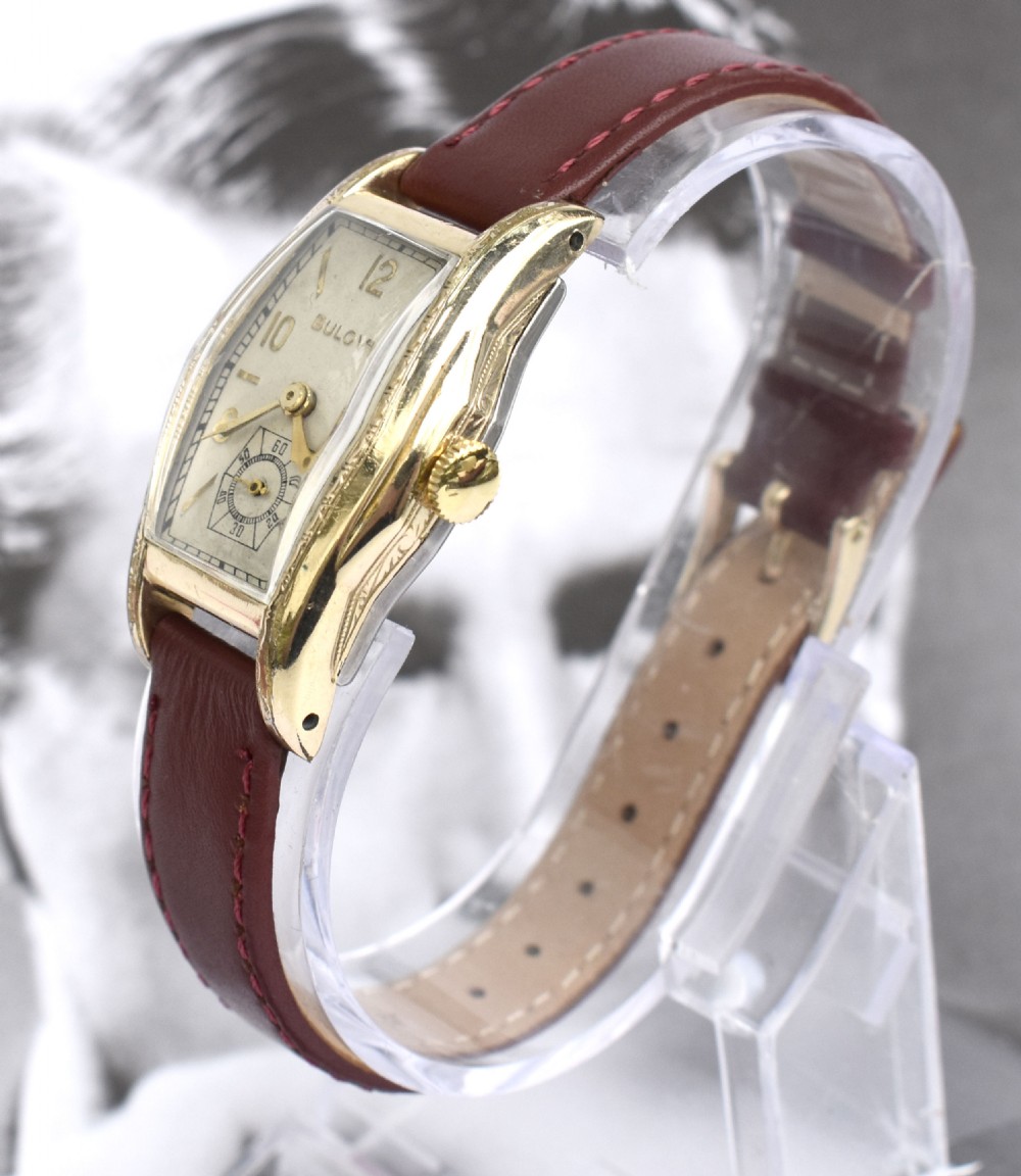 An Art Deco Bulova gents wristwatch featuring a gold-tone tonneau case with engraved side detailing, a cream dial with gold numerals, and a burgundy leather strap. View IX