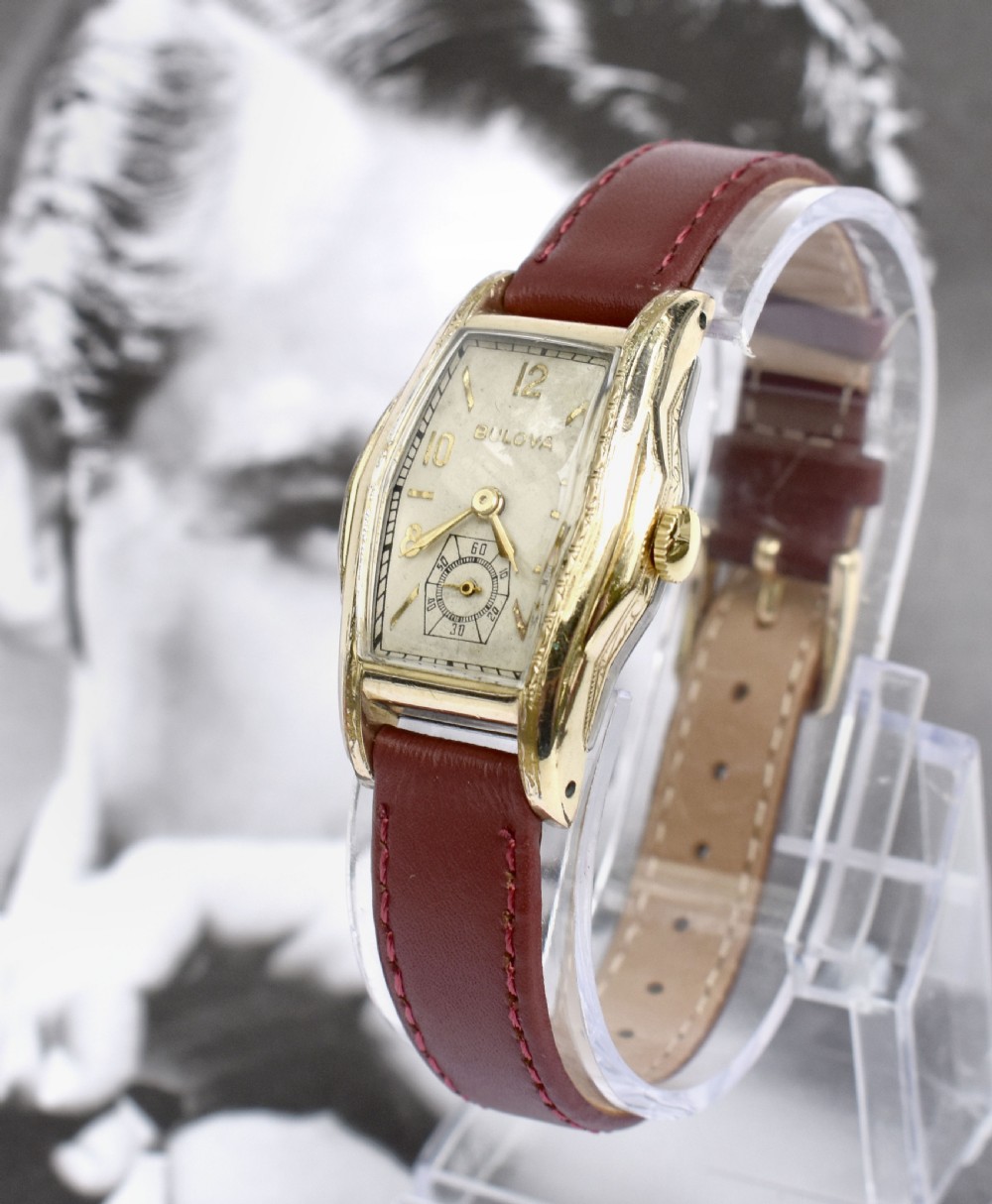 An Art Deco Bulova gents wristwatch featuring a gold-tone tonneau case with engraved side detailing, a cream dial with gold numerals, and a burgundy leather strap. View X