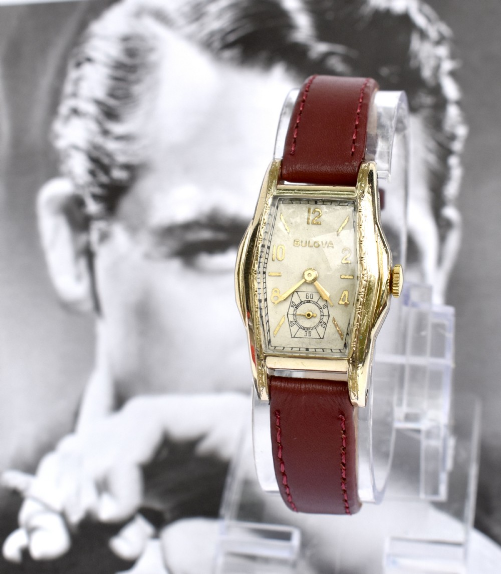 An Art Deco Bulova gents wristwatch featuring a gold-tone tonneau case with engraved side detailing, a cream dial with gold numerals, and a burgundy leather strap. View XI