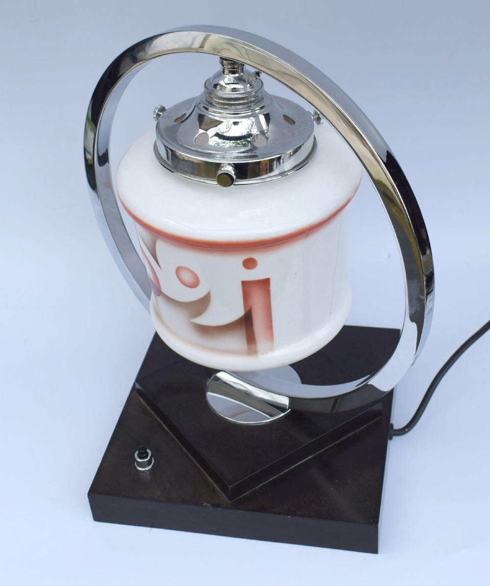 An Art Deco table lamp featuring a circular chrome hoop frame around a patterned glass shade, set on a dark brown stepped Bakelite base. View II