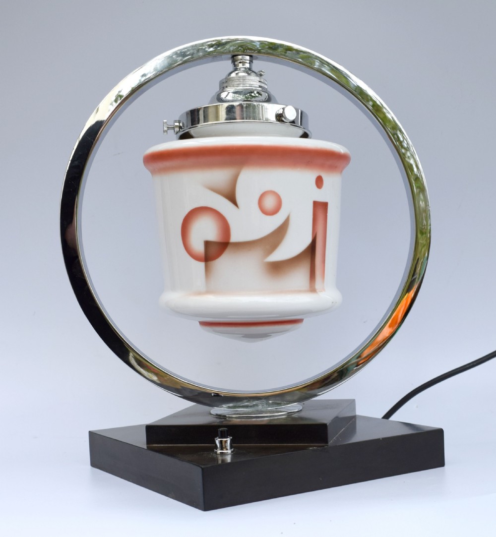 An Art Deco table lamp featuring a circular chrome hoop frame around a patterned glass shade, set on a dark brown stepped Bakelite base. View IV