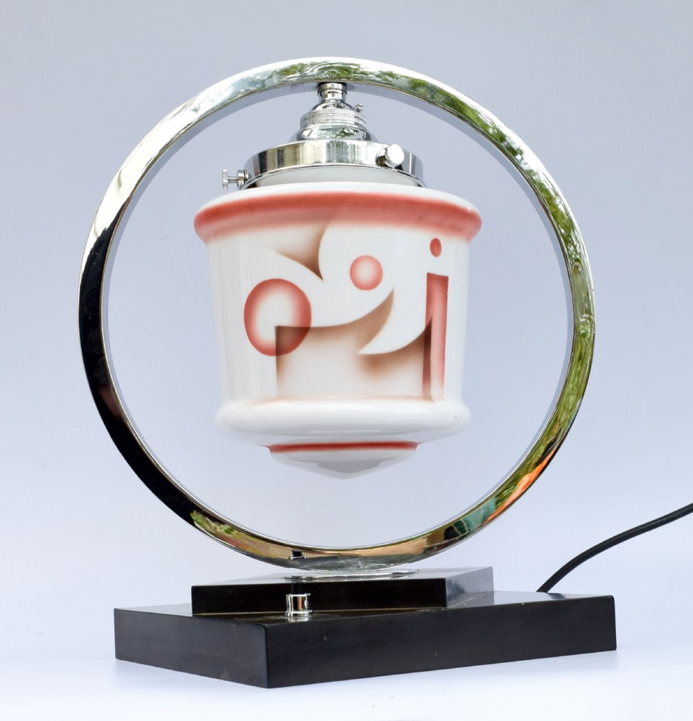 An Art Deco table lamp featuring a circular chrome hoop frame around a patterned glass shade, set on a dark brown stepped Bakelite base. View V