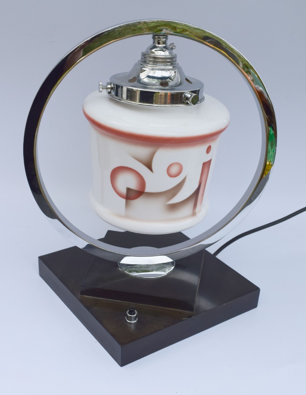 An Art Deco table lamp featuring a circular chrome hoop frame around a patterned glass shade, set on a dark brown stepped Bakelite base. View VI