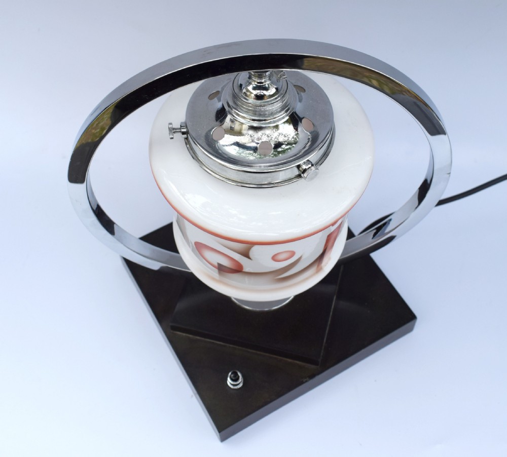 An Art Deco table lamp featuring a circular chrome hoop frame around a patterned glass shade, set on a dark brown stepped Bakelite base. View VII