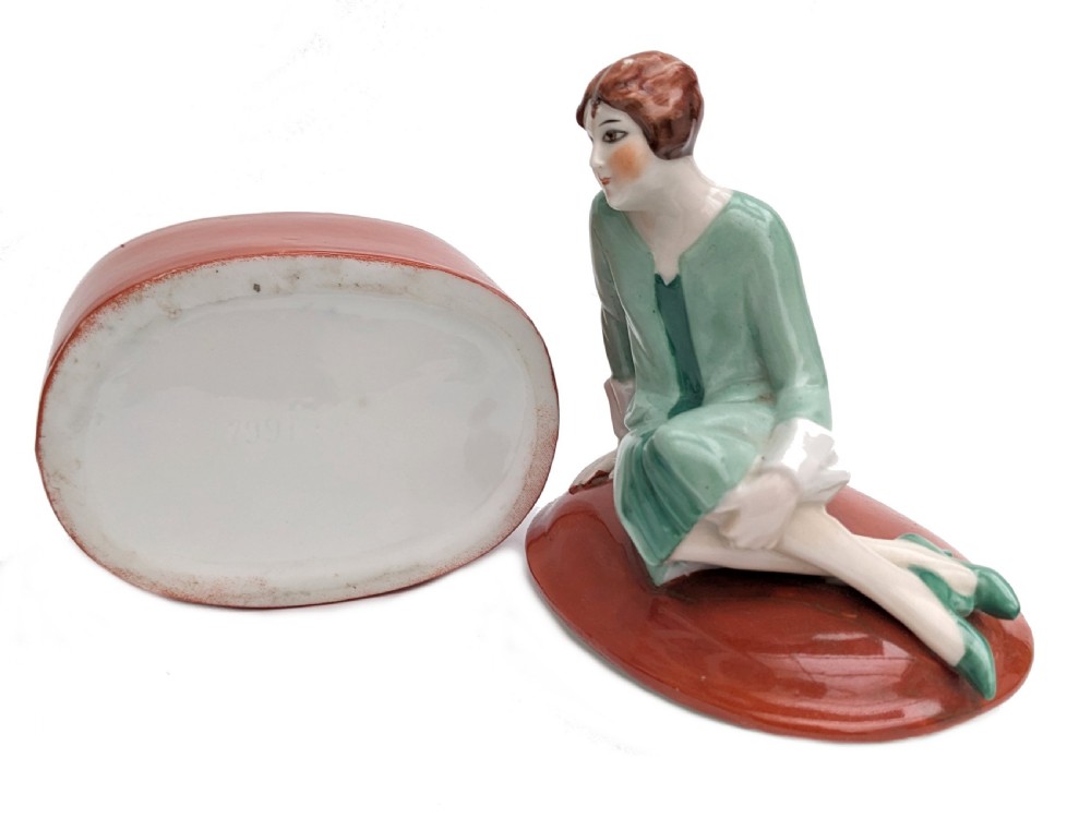 An Art Deco glazed porcelain powder box with a terracotta cylindrical base and a lid surmounted by a painted figurine of a flapper seated in a green dress. View VI