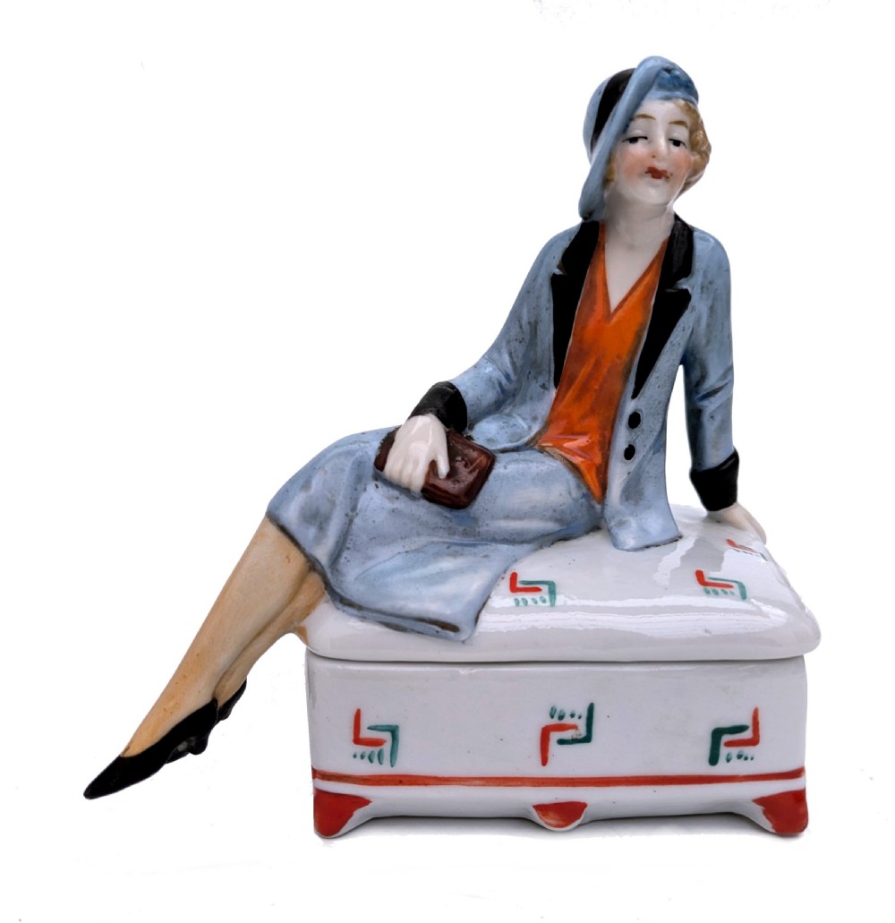 An Art Deco porcelain powder box featuring a sculpted lid shaped as a seated flapper girl dressed in a pale blue suit atop a geometric patterned box.