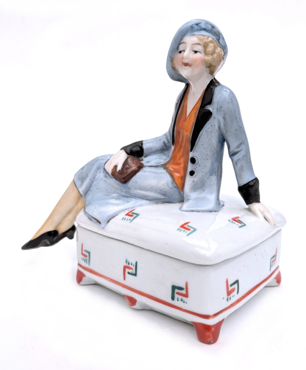 An Art Deco porcelain powder box featuring a sculpted lid shaped as a seated flapper girl dressed in a pale blue suit atop a geometric patterned box. View II