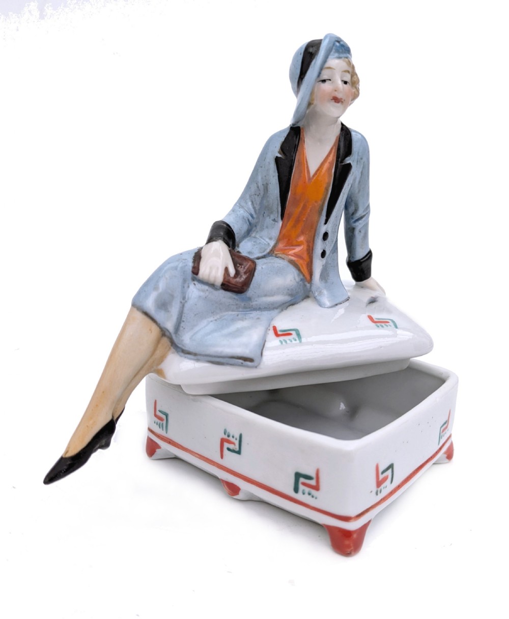 An Art Deco porcelain powder box featuring a sculpted lid shaped as a seated flapper girl dressed in a pale blue suit atop a geometric patterned box. View III