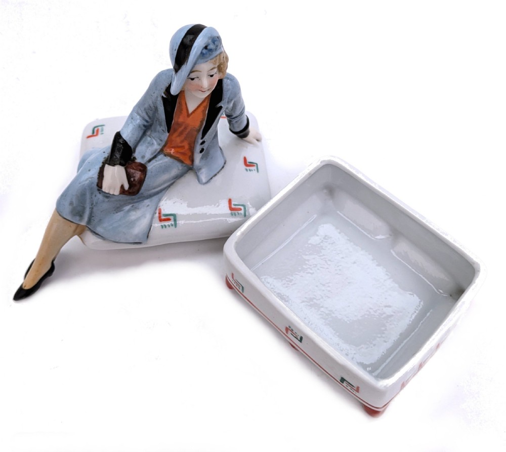 An Art Deco porcelain powder box featuring a sculpted lid shaped as a seated flapper girl dressed in a pale blue suit atop a geometric patterned box. View IV
