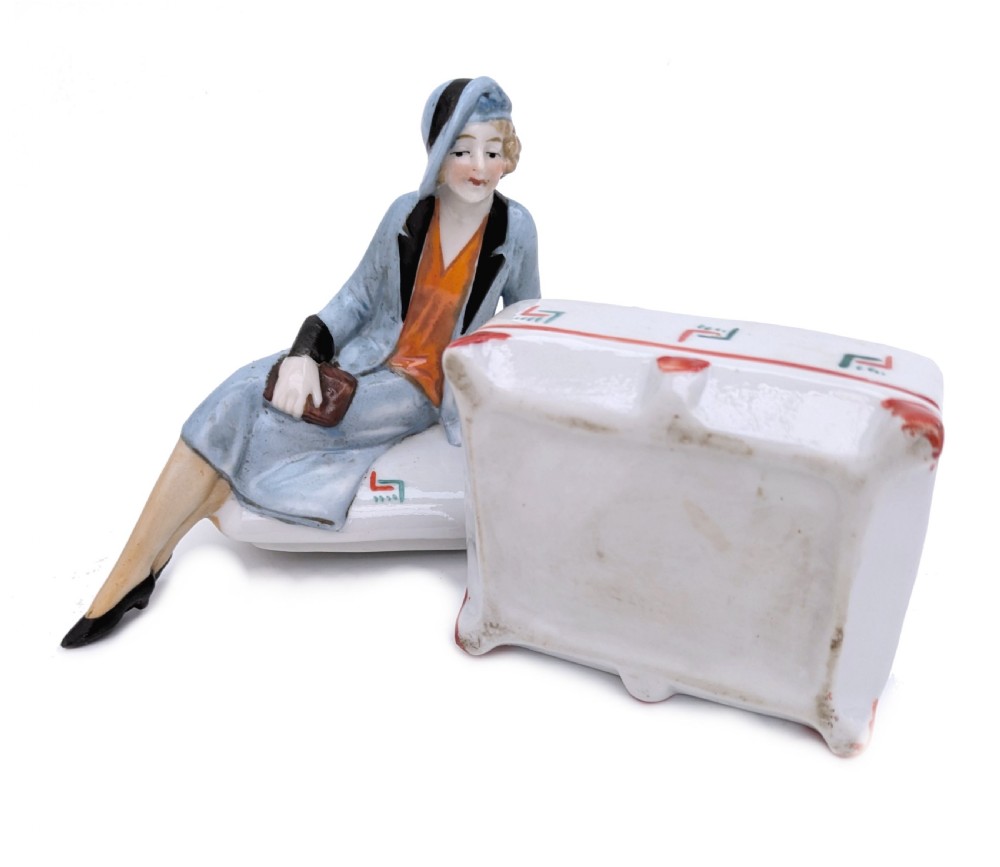 An Art Deco porcelain powder box featuring a sculpted lid shaped as a seated flapper girl dressed in a pale blue suit atop a geometric patterned box. View V