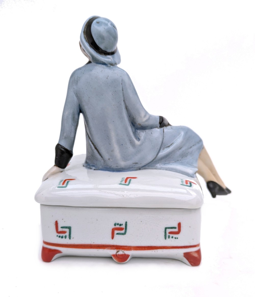 An Art Deco porcelain powder box featuring a sculpted lid shaped as a seated flapper girl dressed in a pale blue suit atop a geometric patterned box. View VI