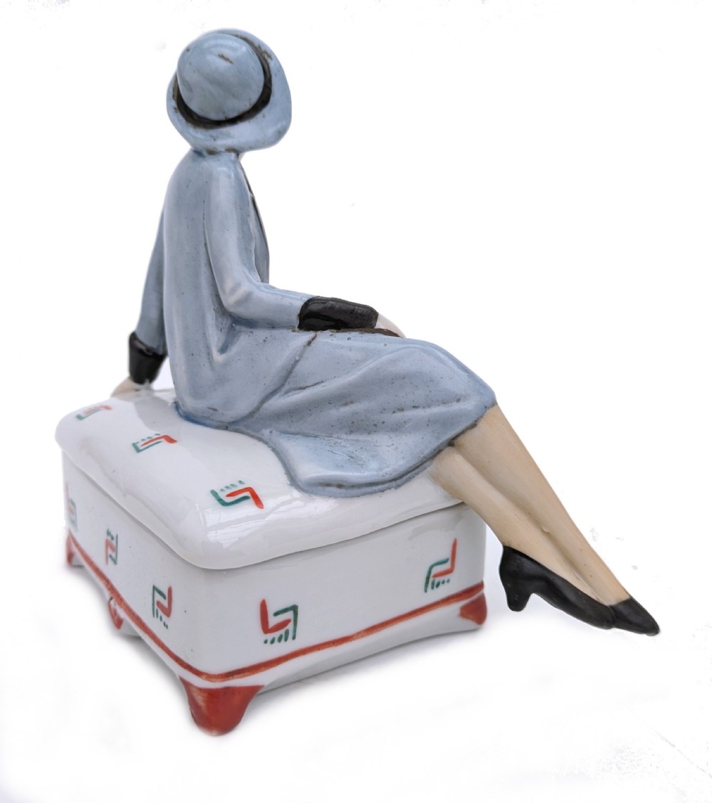 An Art Deco porcelain powder box featuring a sculpted lid shaped as a seated flapper girl dressed in a pale blue suit atop a geometric patterned box. View VII