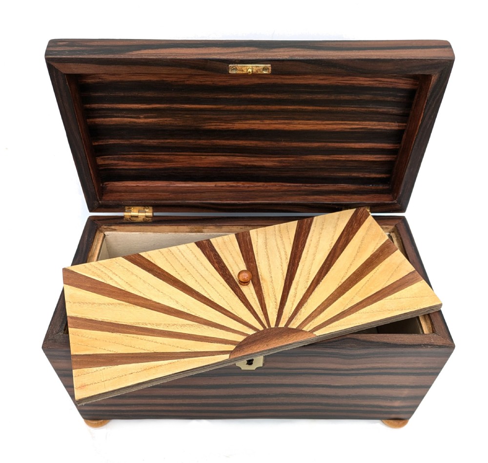 An open Art Deco dark wood tea caddy featuring a light wood sunburst inlay pattern on its internal lid.