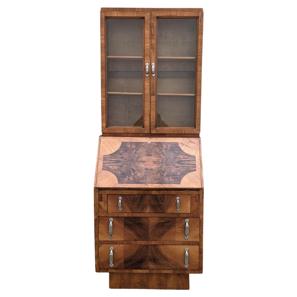 An Art Deco walnut upright bureau bookcase featuring a two-door glazed upper display cabinet above a slope-front writing slope and three drawers.