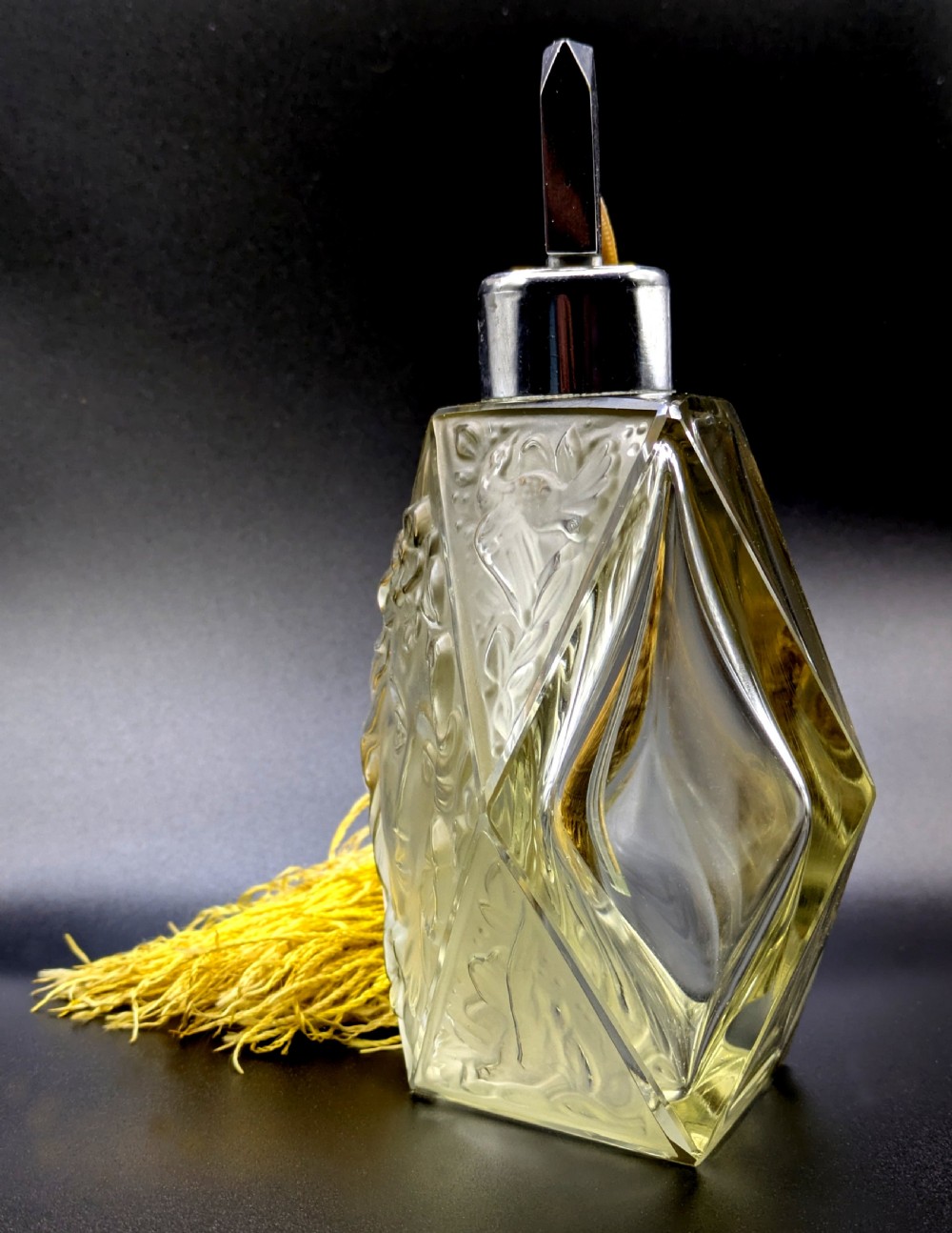 A faceted clear glass Art Deco scent bottle featuring a relief-moulded womans face and a gold fabric atomiser bulb with tassel. View IV