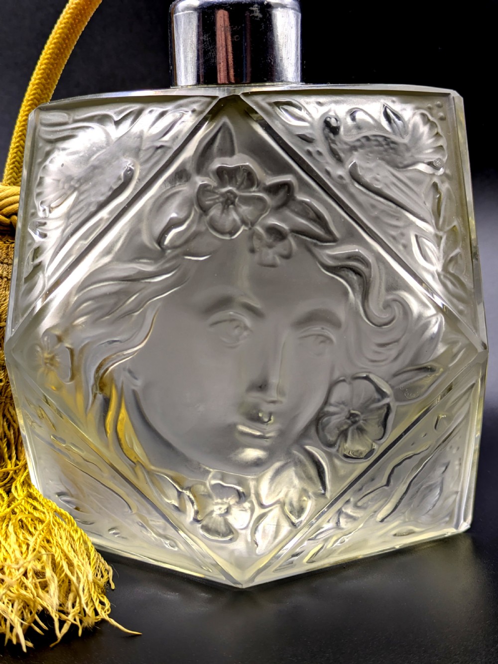 A faceted clear glass Art Deco scent bottle featuring a relief-moulded womans face and a gold fabric atomiser bulb with tassel. View VIII