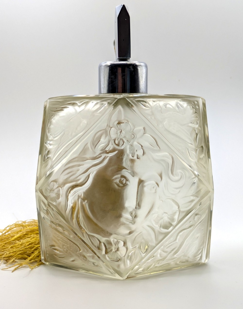 A faceted clear glass Art Deco scent bottle featuring a relief-moulded womans face and a gold fabric atomiser bulb with tassel. View XVI