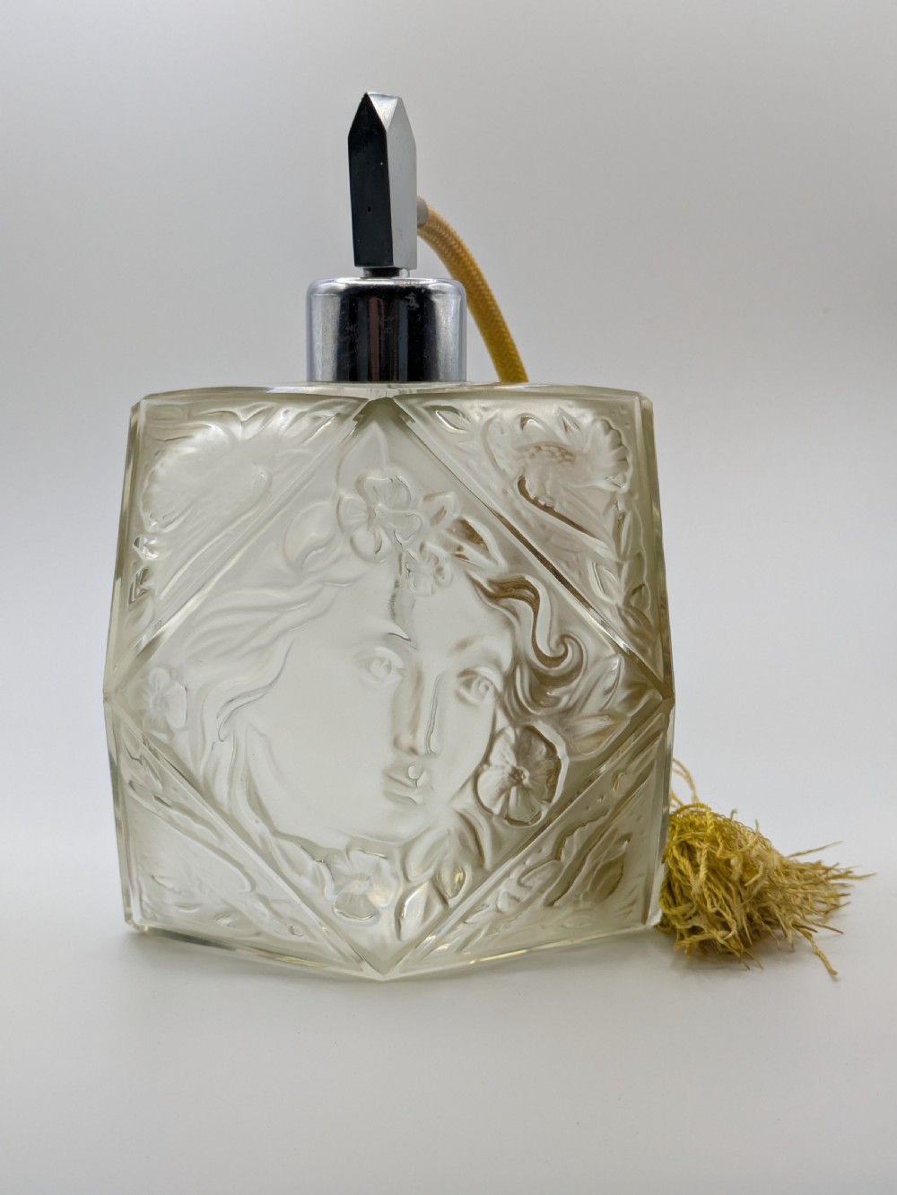 A faceted clear glass Art Deco scent bottle featuring a relief-moulded womans face and a gold fabric atomiser bulb with tassel. View XX