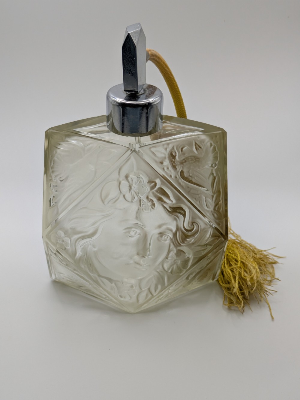 A faceted clear glass Art Deco scent bottle featuring a relief-moulded womans face and a gold fabric atomiser bulb with tassel. View XXI