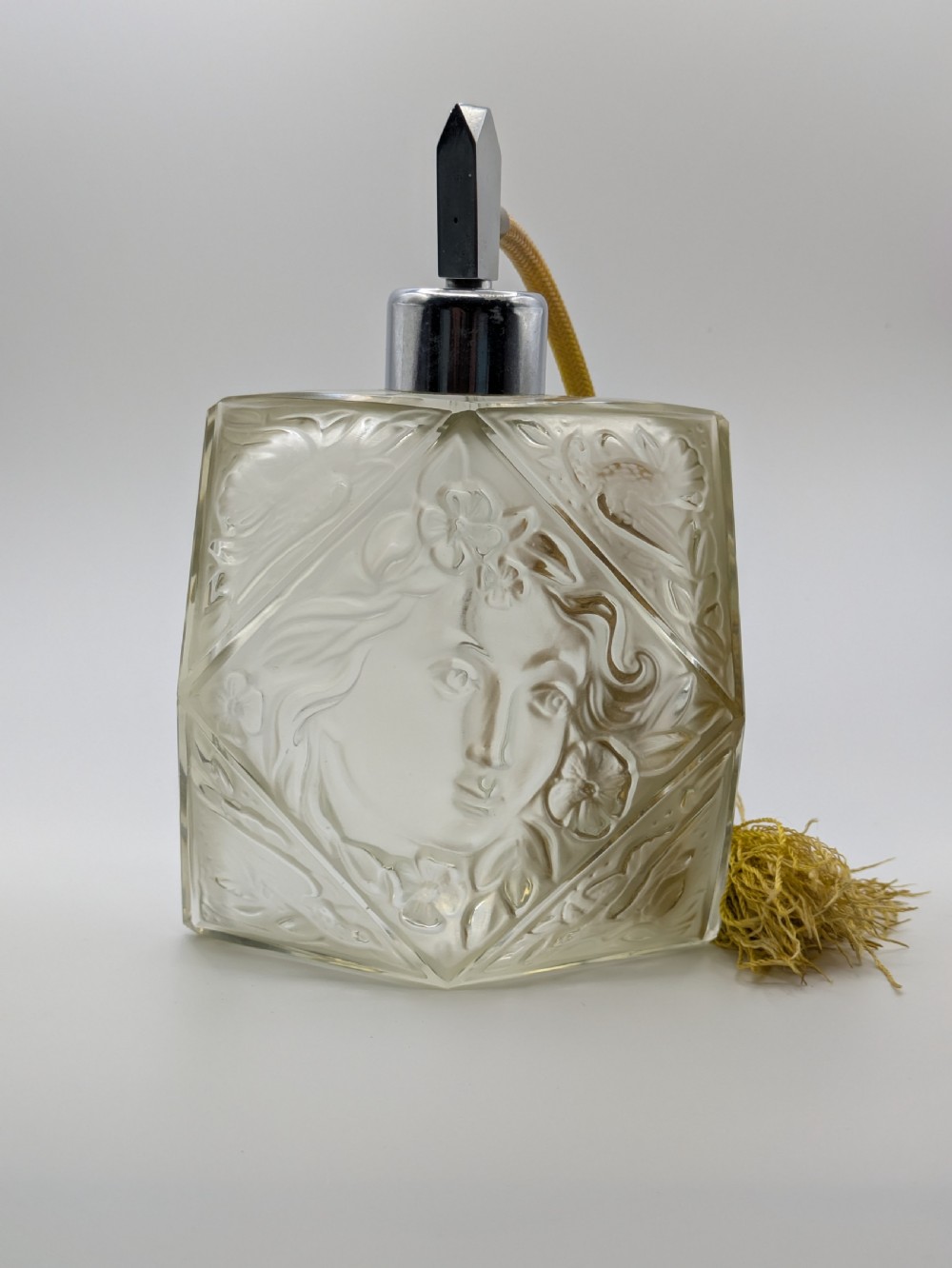 A faceted clear glass Art Deco scent bottle featuring a relief-moulded womans face and a gold fabric atomiser bulb with tassel. View XXII