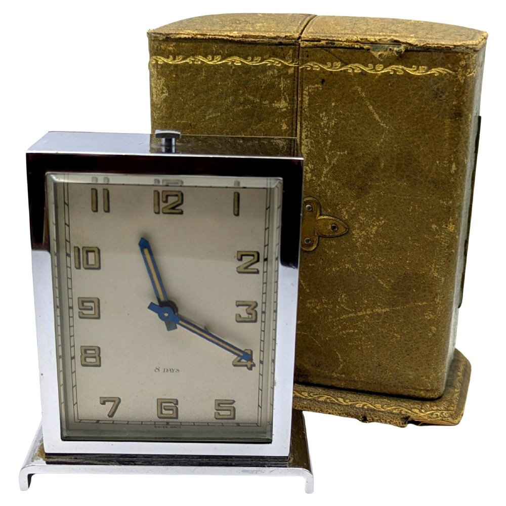 A rectangular Art Deco chrome-plated travel clock with blue hands and geometric numerals beside its fitted tan leather case.