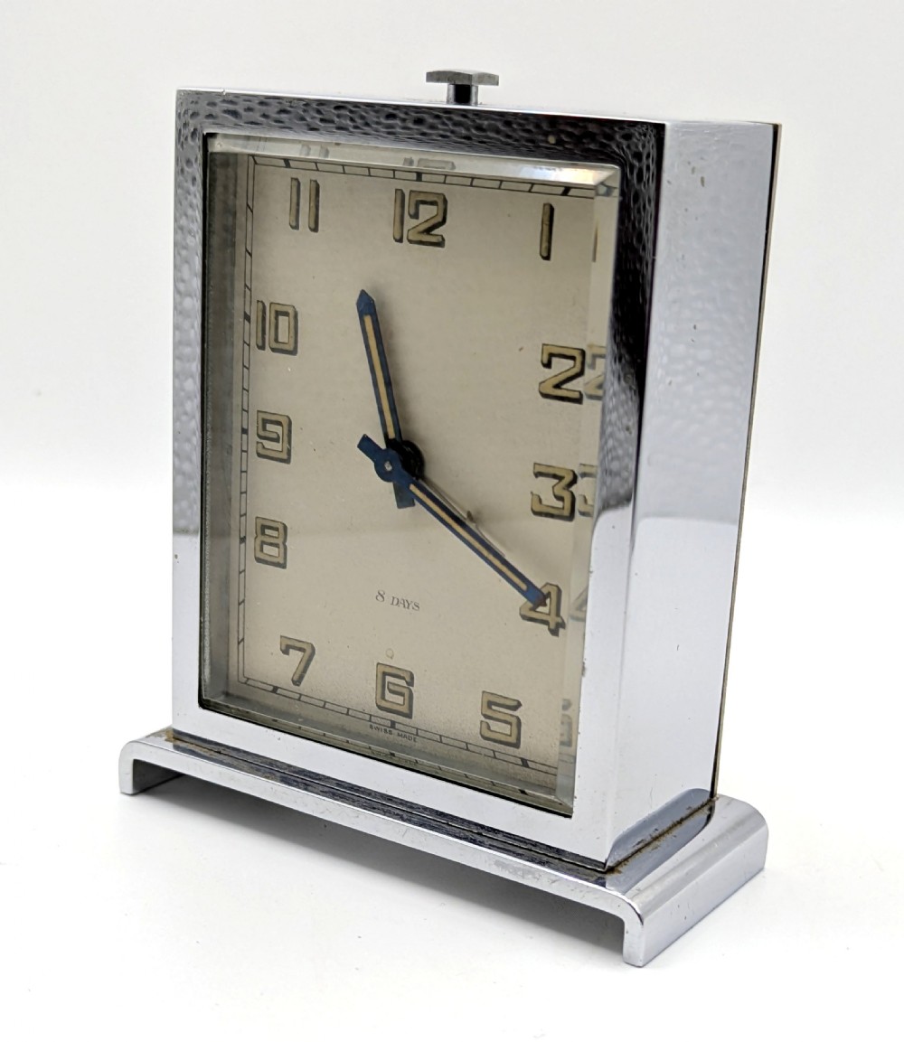 A rectangular Art Deco chrome-plated travel clock with blue hands and geometric numerals beside its fitted tan leather case. View VII