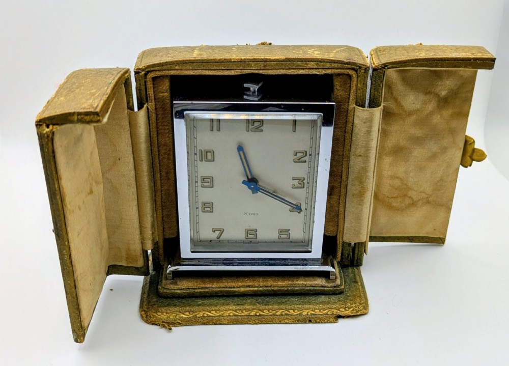 A rectangular Art Deco chrome-plated travel clock with blue hands and geometric numerals beside its fitted tan leather case. View IX